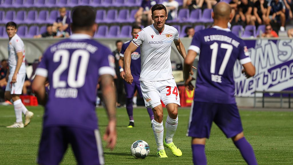 Fußballspiel auf einem grünen Platz. Einige Spieler tragen weiße Kleidung. Andere tragen dunkle Kleidung. Im Hintergrund stehen Zuschauer.