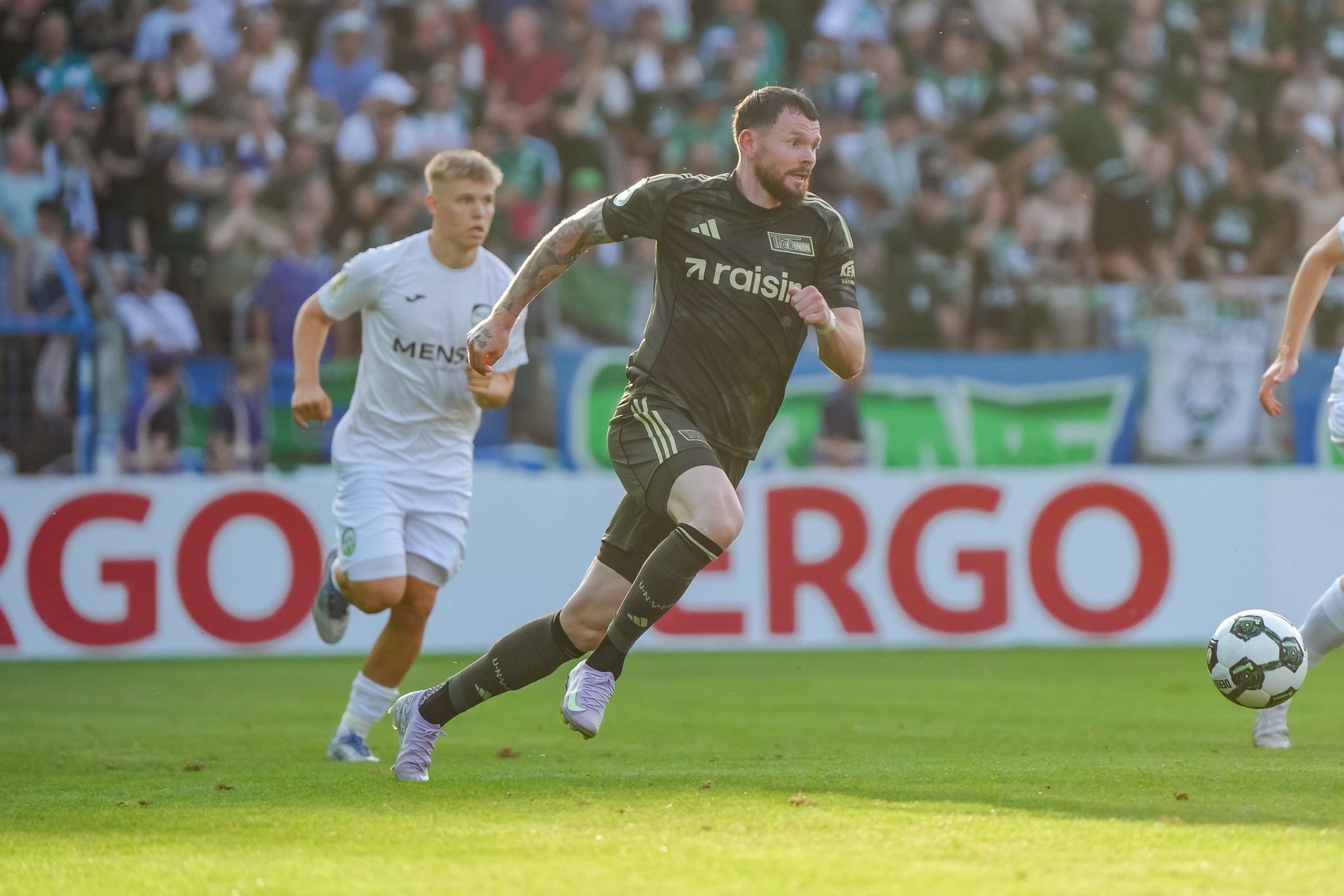 Ein Spieler in schwarzem Trikot sprintet mit dem Ball, während ein Gegner in weißem Trikot ihn verfolgt. Hintergrund mit jubelnden Fans.