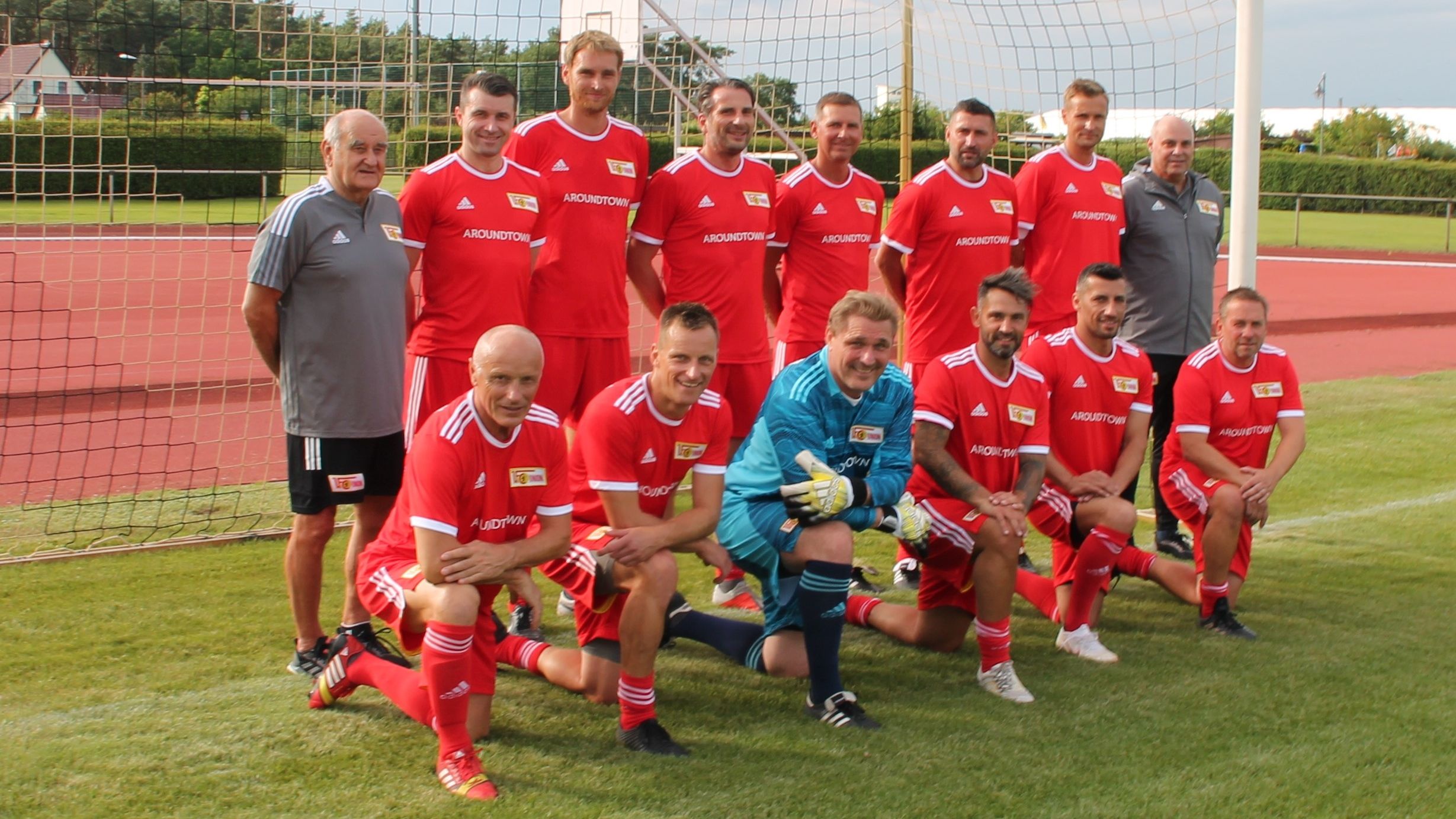 Eine Fußballmannschaft in roten Trikots posiert auf einem Platz, umgeben von einem Tor und grünen Rasen.