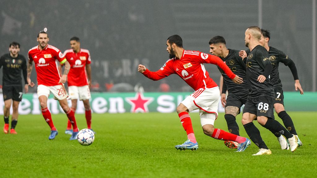 Ein Fußballspieler in rotem Trikot sprintet mit dem Ball, während zwei Verteidiger in schwarzen Trikots ihn verfolgen.