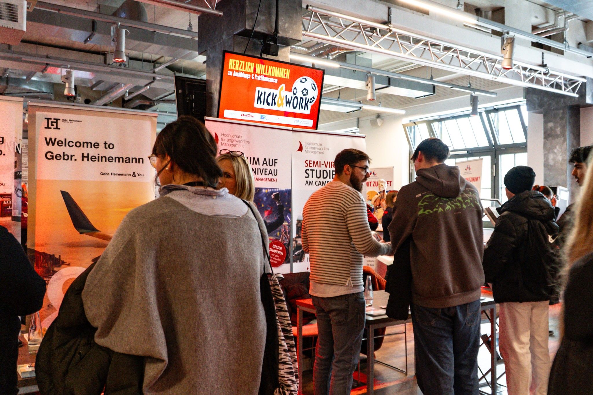 Personen stehen an Messeständen in einer Veranstaltungshalle, während ein Bildschirm Informationen und Begrüßungen anzeigt.