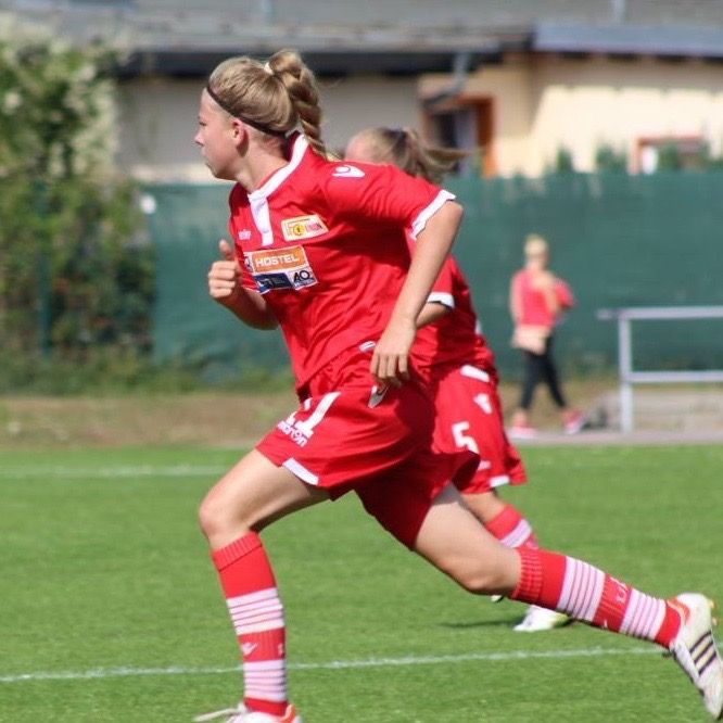 Eine junge Frau in roter Fußballausrüstung sprintet auf einem Rasenfeld, während weitere Spieler im Hintergrund stehen.
