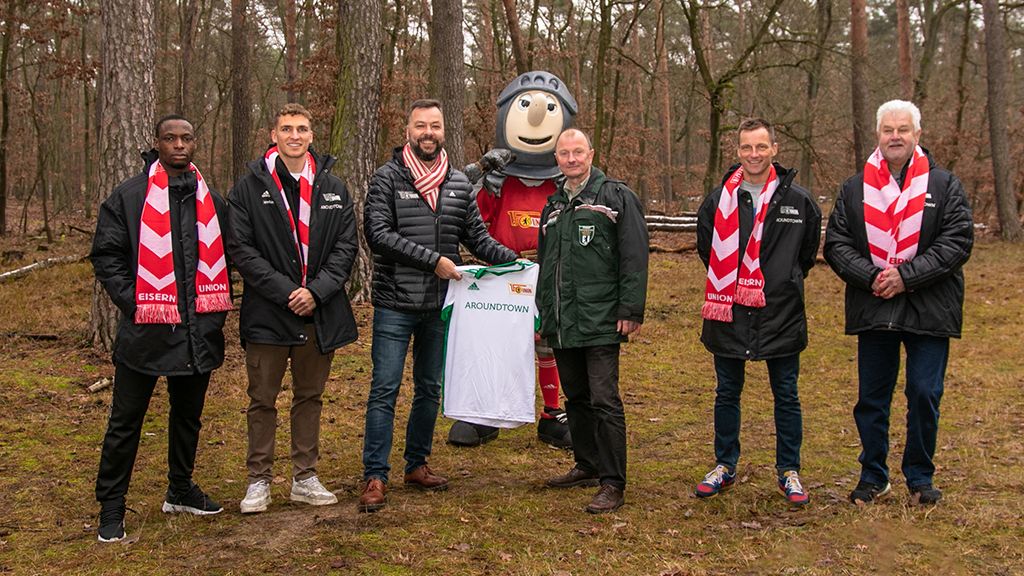 Sechs Personen stehen in einem Wald, tragen Schals und halten ein Trikot mit der Aufschrift "AROUND TOWN". Ein Maskottchen ist ebenfalls dabei.