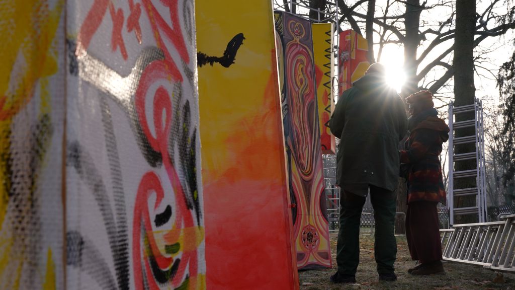Zwei Menschen stehen zwischen bunten, bemalten Panels, während die Sonne im Hintergrund aufgeht.