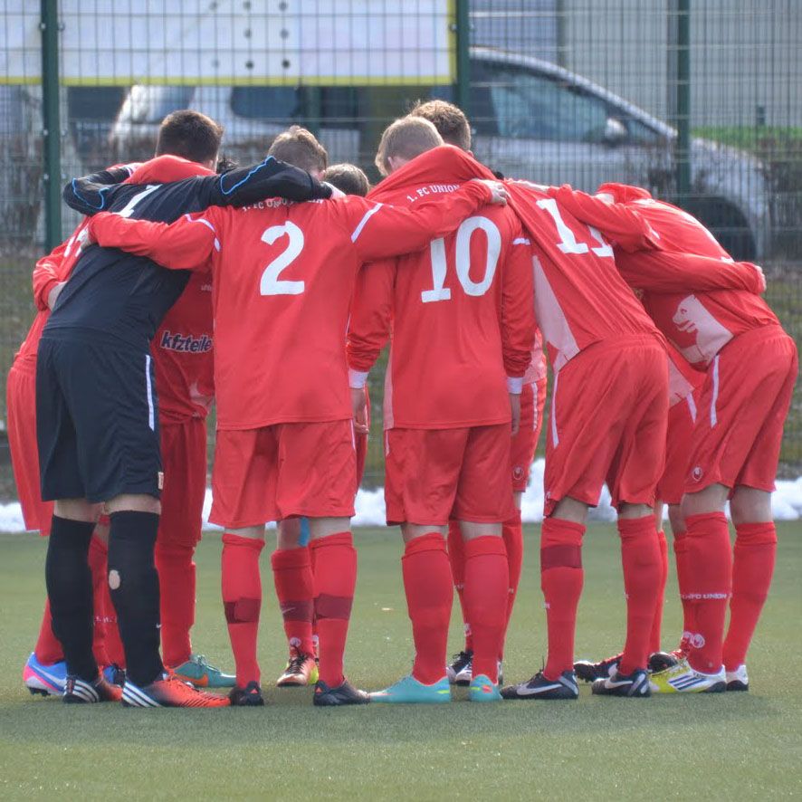 Spieler in roten Fußballtrikots stehen eng zusammen und bilden einen Kreis auf dem Spielfeld.