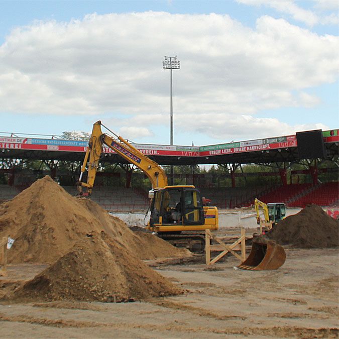 Bauarbeiten im Stadion mit Baggern und Sandhaufen unter einem bewölkten Himmel.