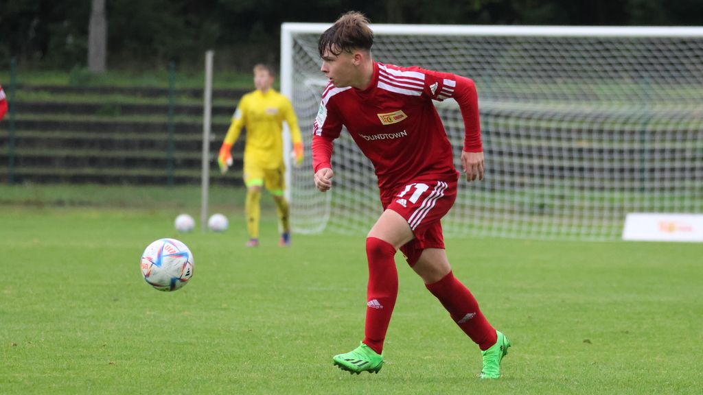 Ein junger Fußballspieler in rotem Trikot dribbelt den Ball auf einem Rasenplatz. Im Hintergrund steht ein Torwart.