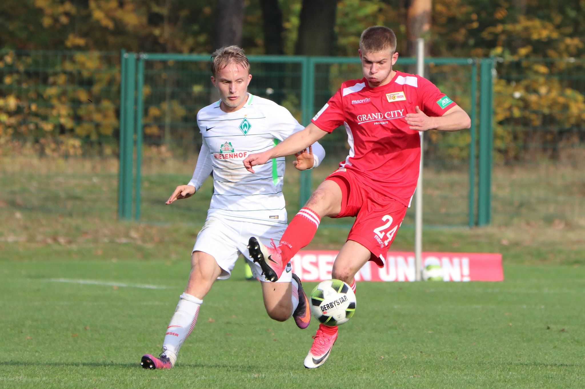 Two football players in action, one in red and the other in white, on a grassy field with trees in the background.