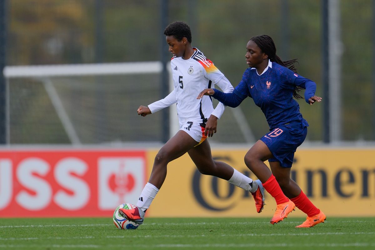 Zwei Fußballspielerinnen im Wettkampf auf einem Spielfeld, eine trägt ein weißes Trikot mit schwarzer Nummer, die andere ein blaues Trikot.
