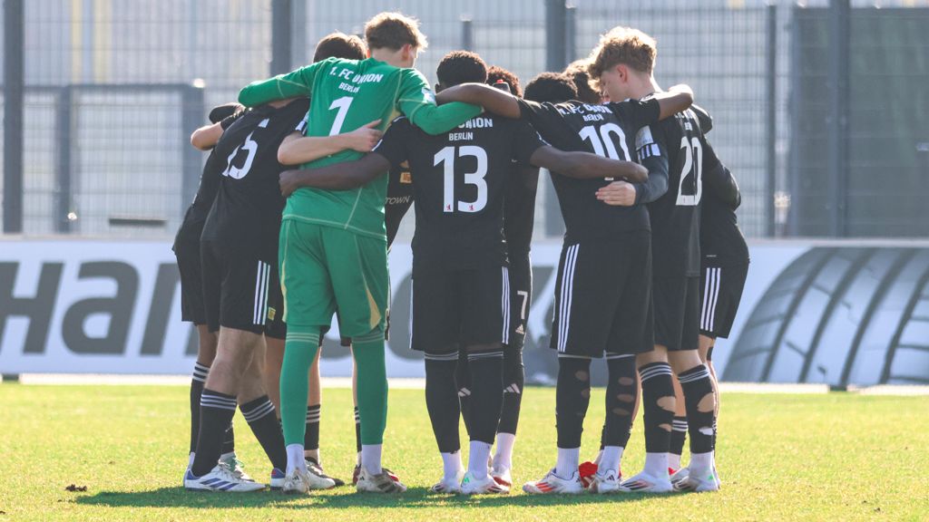 Eine Gruppe von Fußballspielern in schwarzen Trikots umarmt sich im Kreis auf einem Fußballfeld.