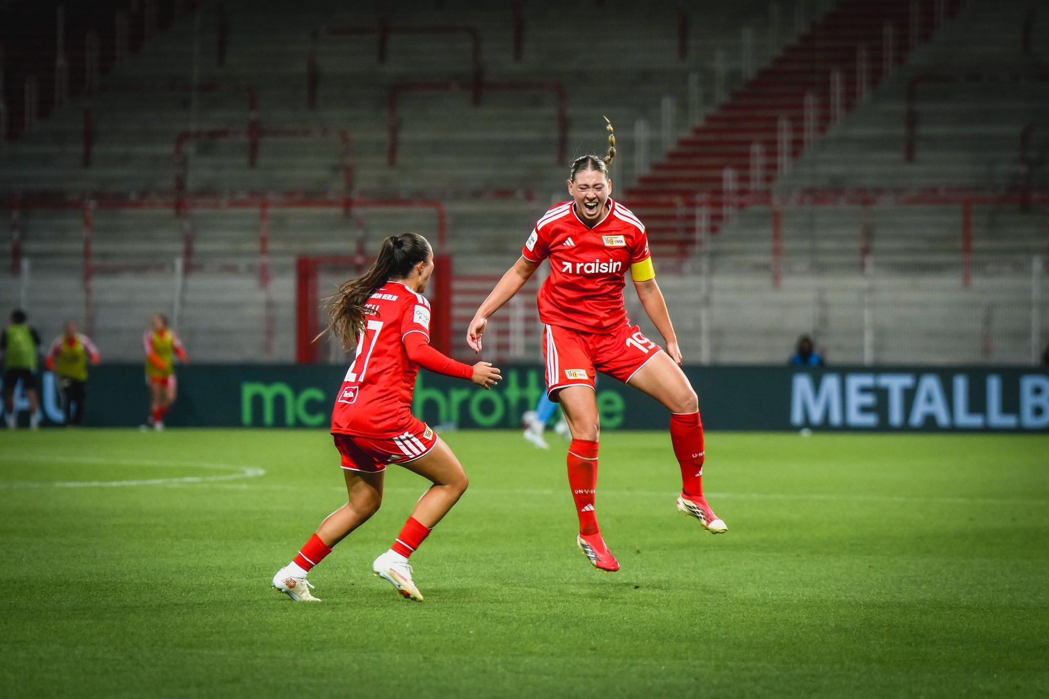 Zwei Fußballspielerinnen in roten Trikots feiern während eines Spiels auf dem Spielfeld.