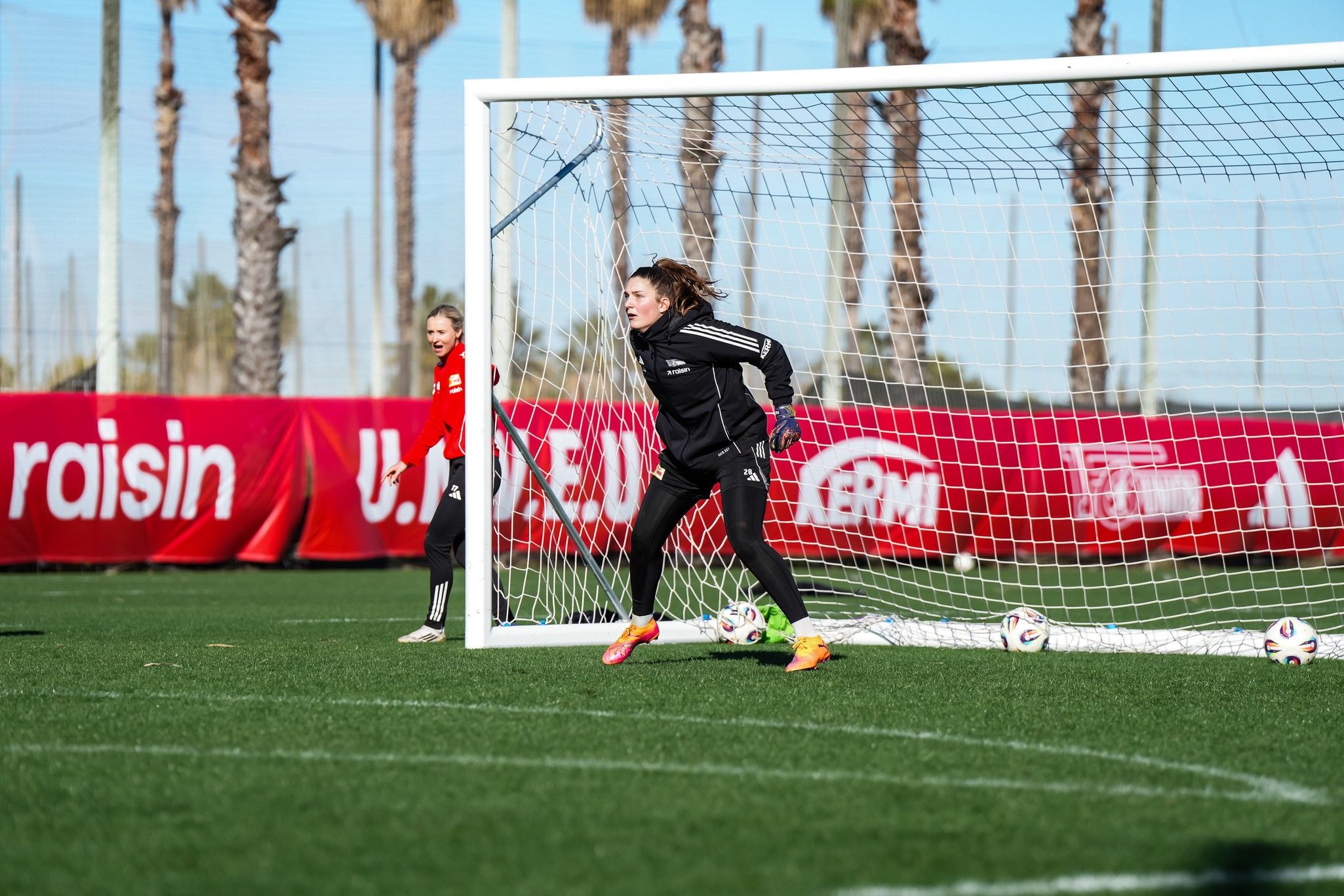 Torhüterin in einem Fußballtor während des Trainings, bereit zum Abfangen eines Schusses. Im Hintergrund Palmen und Trainingskollegen.
