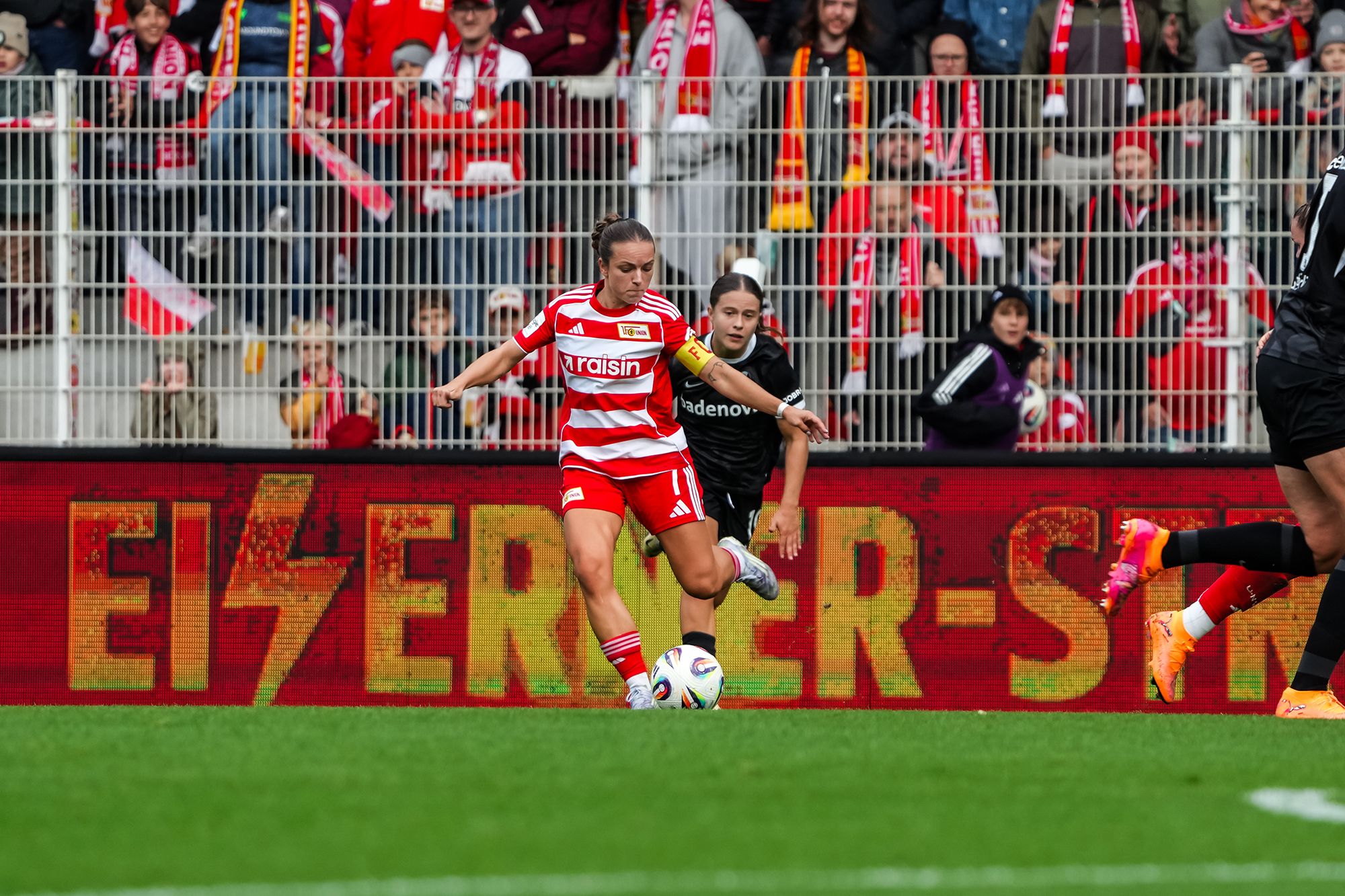 Eine Spielerin in rotem Trikot dribbelt den Ball, während eine Torhüterin und Zuschauer im Hintergrund zu sehen sind.