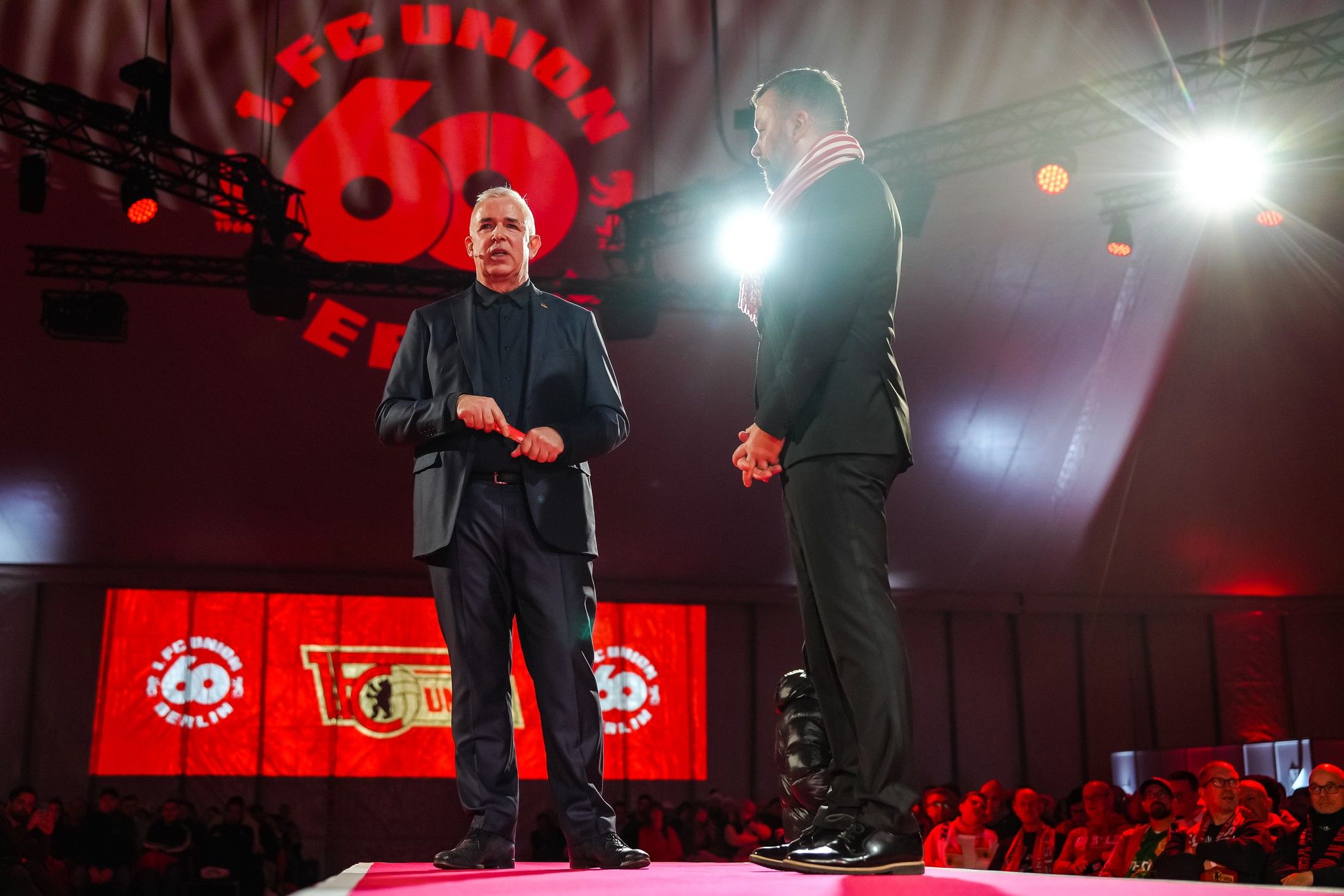 Zwei Männer stehen auf einer Bühne während einer Veranstaltung von 1. FC Union Berlin, umgeben von roten Lichtern und Zuschauern.