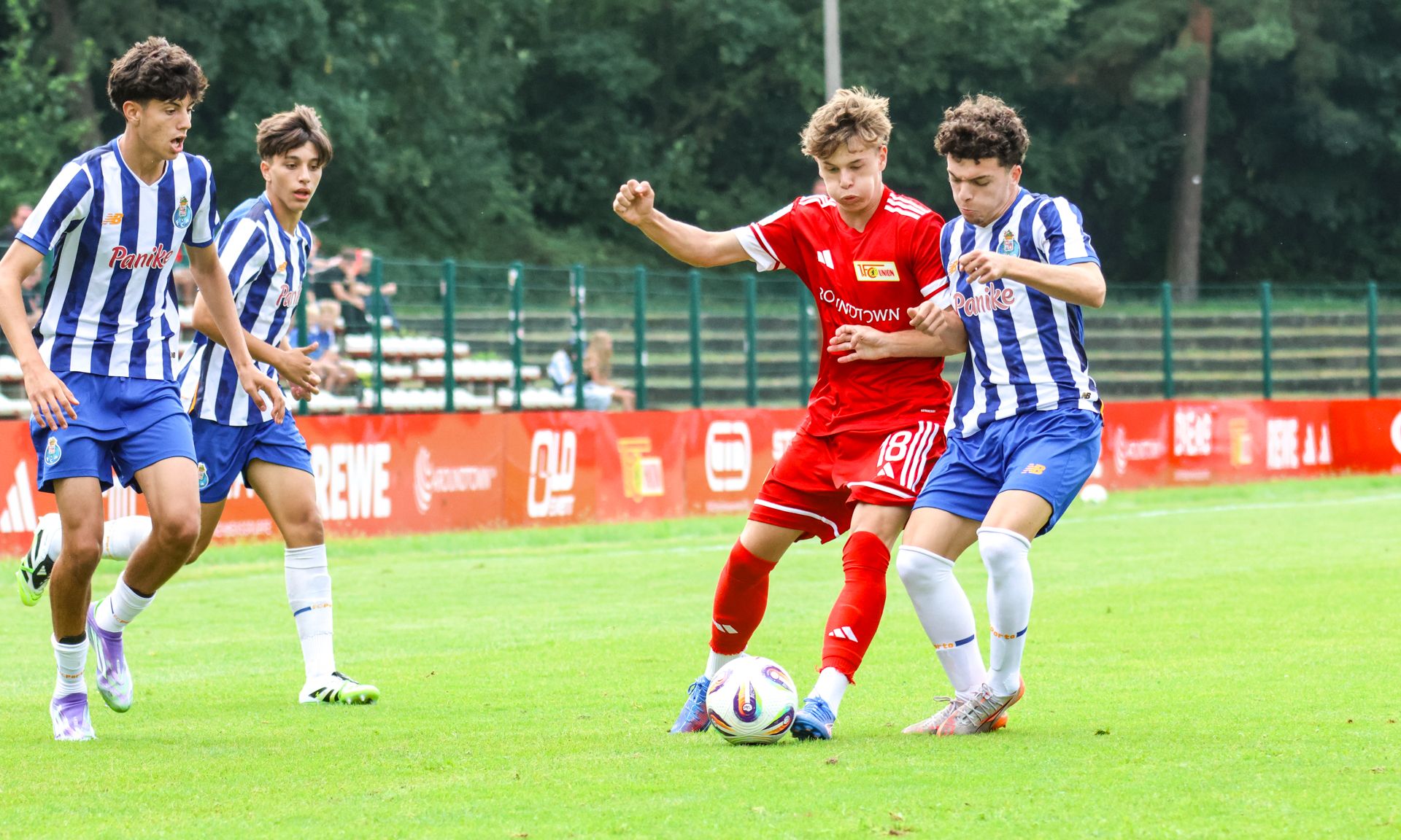 Zwei Fußballspieler kämpfen um den Ball, während zwei weitere Spieler im Hintergrund zuschauen. Das Spiel findet auf einem grünen Platz statt.