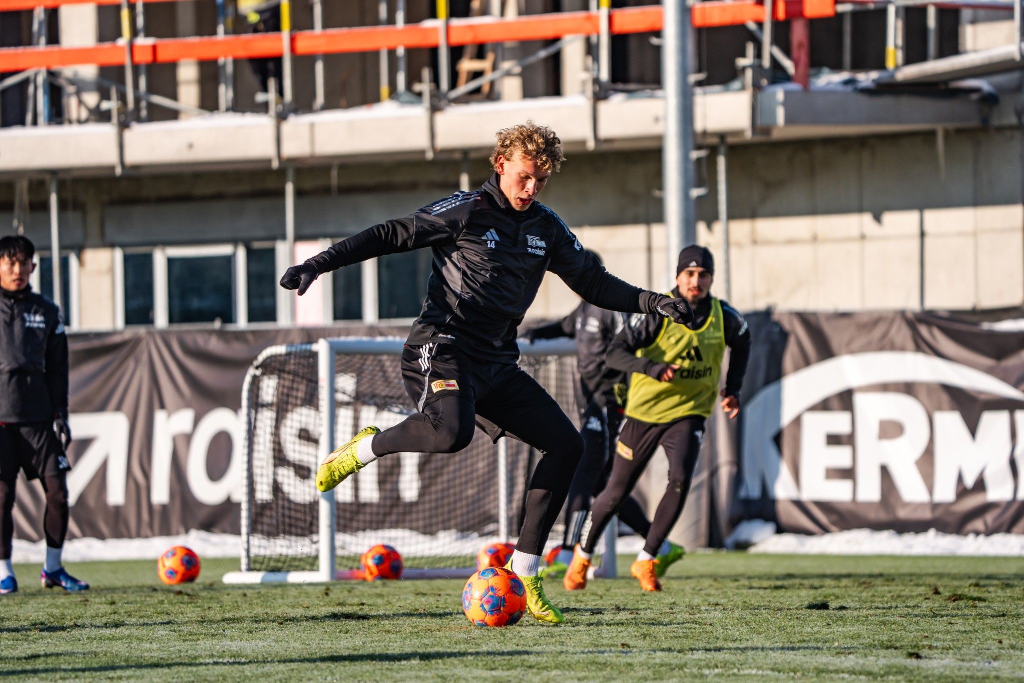 Fußballspieler in Schwarz tritt einen Ball während eines Trainings auf einem Rasenfeld, im Hintergrund Gestalten und Tore.