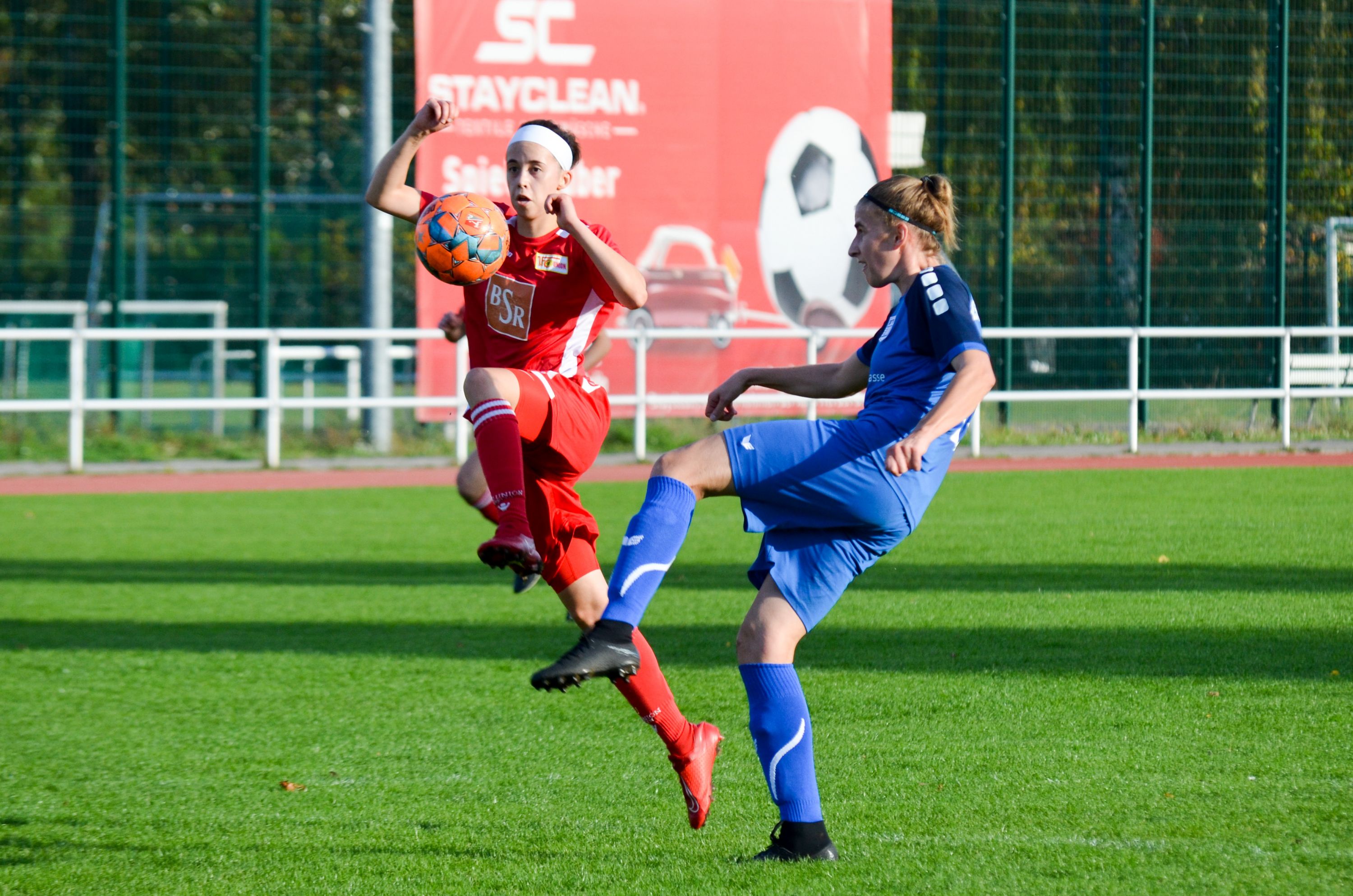 Zwei Fußballspielerinnen spielen auf einem Rasenplatz. Eine trägt Rot, die andere trägt Blau. Sie kämpfen um den Ball.