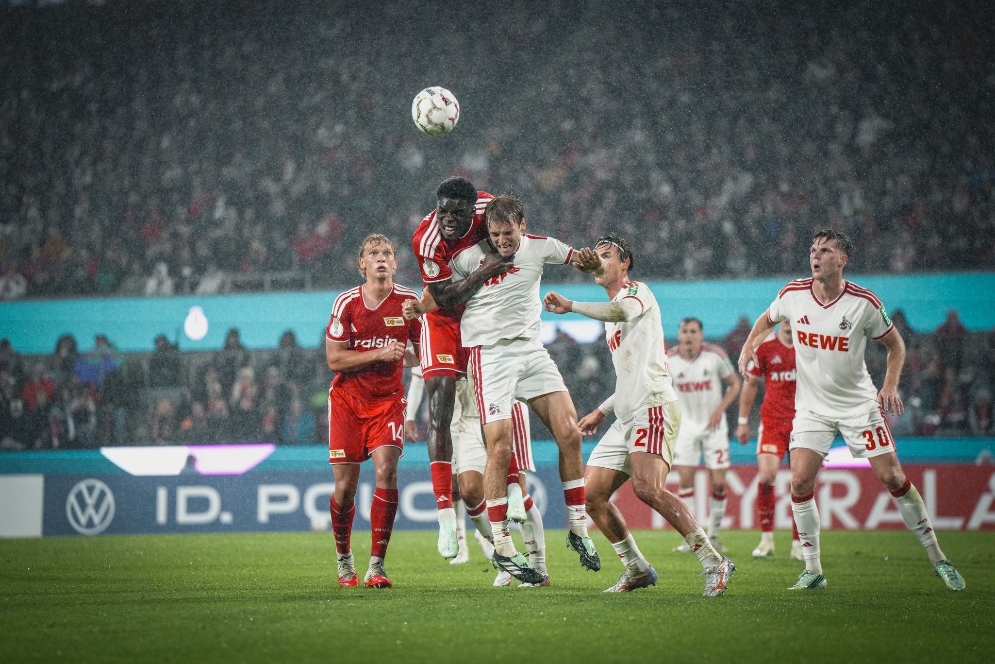 Zwei Fußballspieler springen im Regen hoch, während andere Spieler in der Nähe stehen und das Geschehen beobachten.