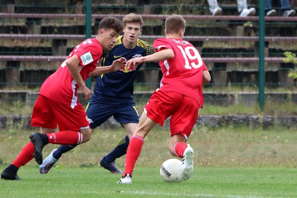 Drei junge Fußballspieler auf dem Platz; zwei in roten Trikots, einer in blauem Trikot, im Moment des Spiels.