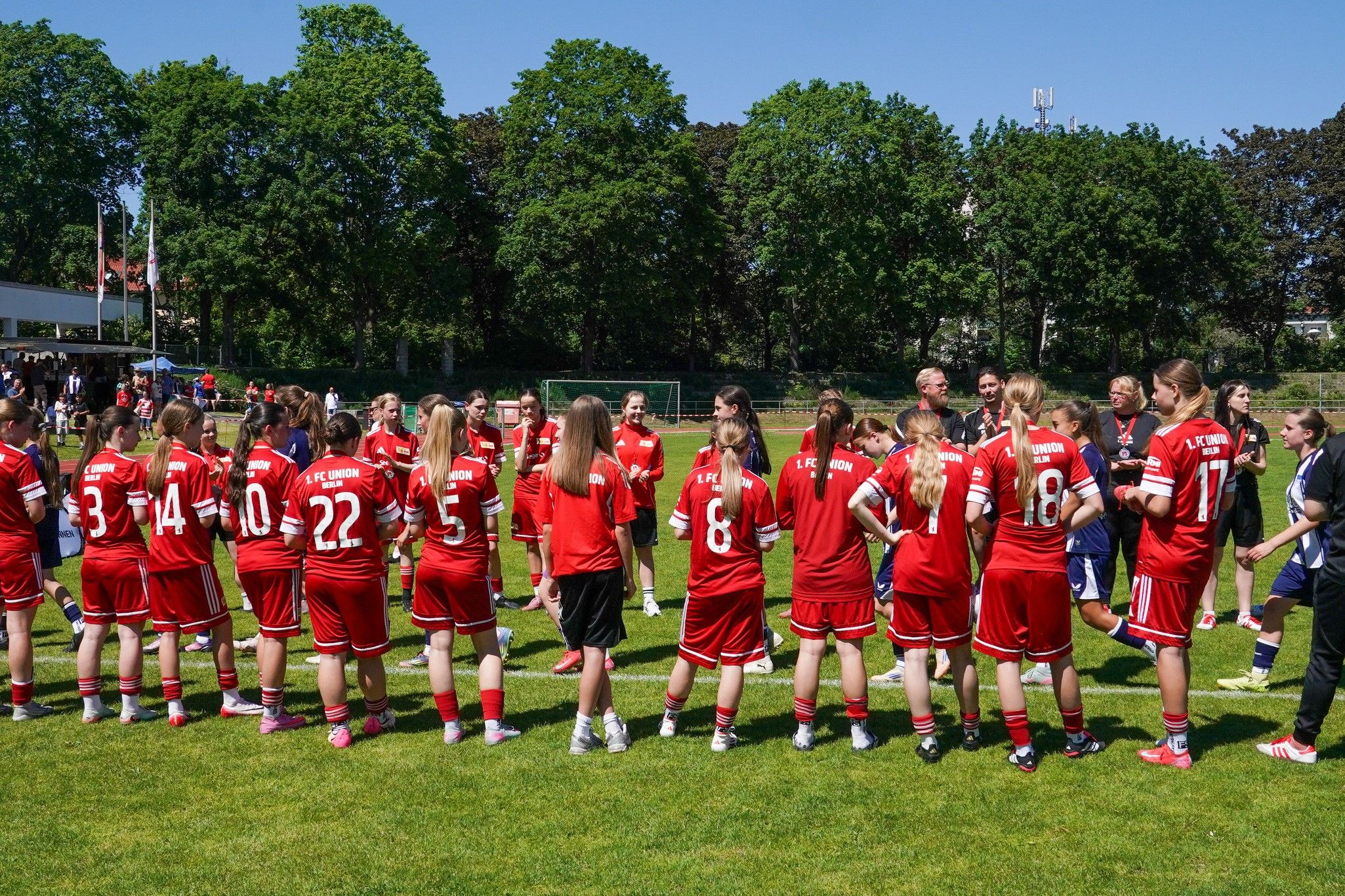 Eine Gruppe von Fußballspielerinnen in roten Trikots steht auf einem Rasenplatz und spricht mit mehreren Erwachsenen.