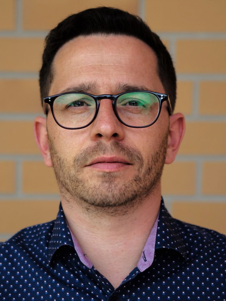 Ein Mann mit Brille und Bart, in einem gepunkteten Hemd, steht vor einer ziegelfarbenen Wand und schaut direkt in die Kamera.