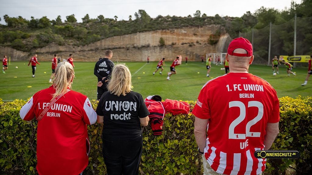 Three football fans in club gear watch a training session of 1. FC Union Berlin, surrounded by a green hedge.