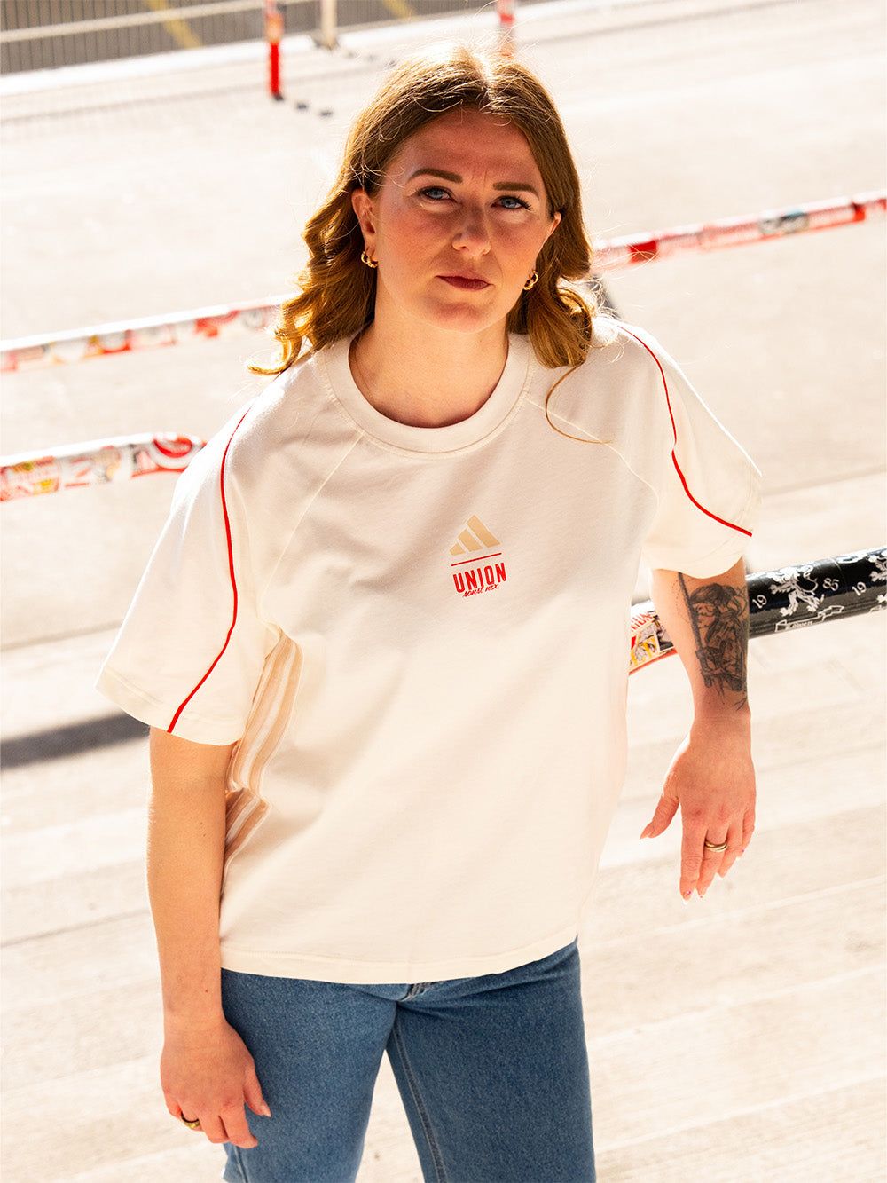 Junge Frau mit langen lockigen Haaren, die in einem cremefarbenen T-Shirt und Jeans an einem Geländer steht.