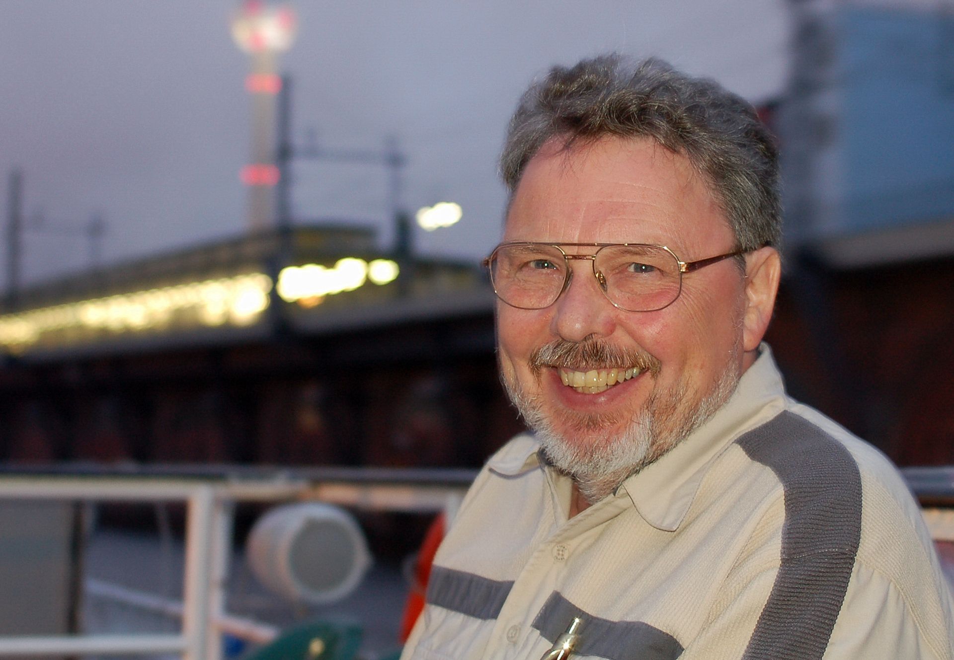 Ein lächelnder Mann mit Brille sitzt auf einem Schiff, im Hintergrund sind Lichter und eine Stadtlandschaft zu sehen.