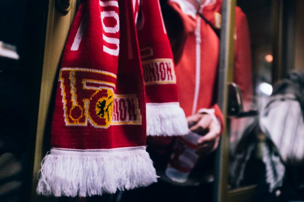 Roter Fan-Schal mit Fransen hängt an einer Tür, im Hintergrund ist eine Person mit rotem Oberteil sichtbar.