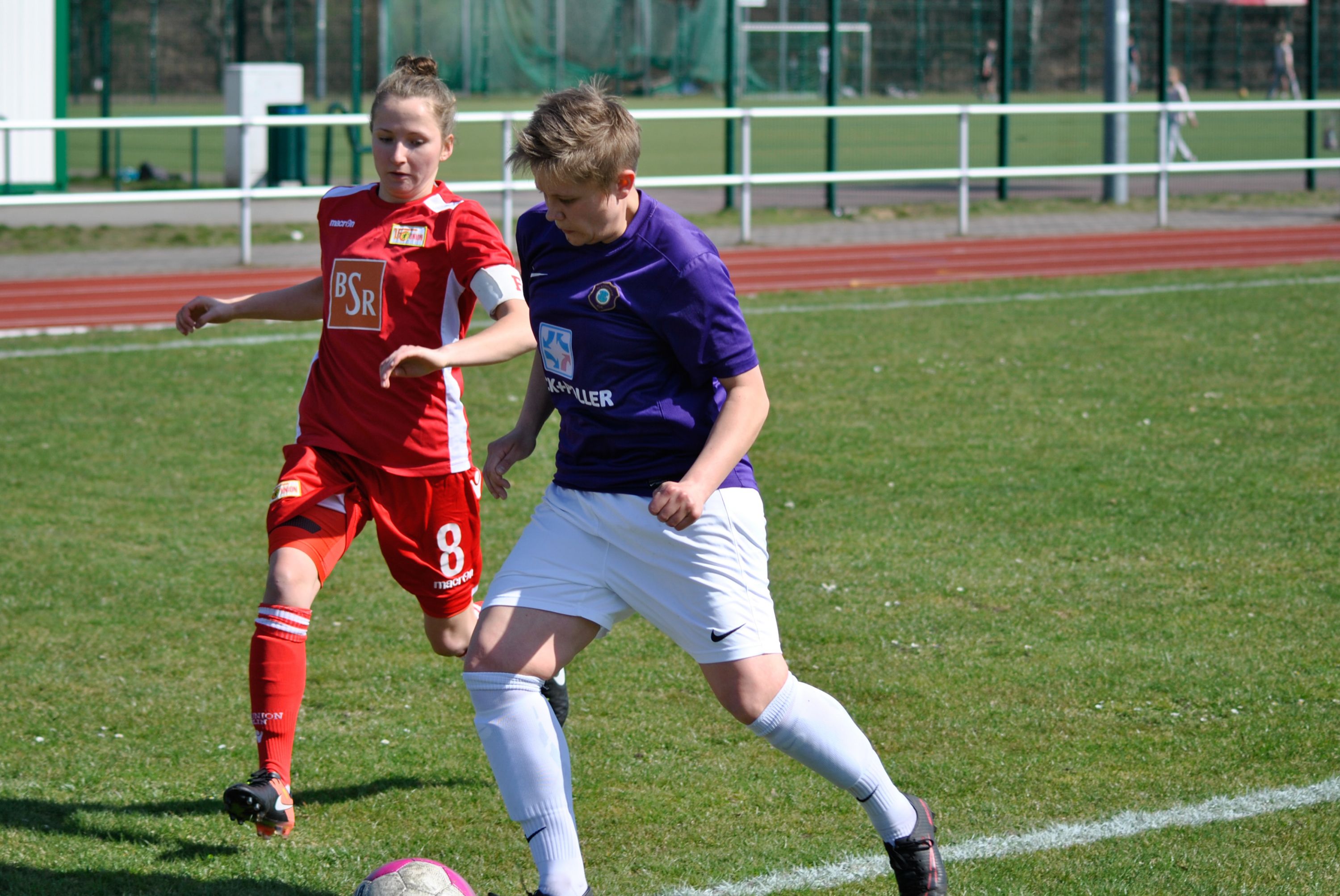 Zwei Frauen spielen Fußball. Sie sind auf einem grünen Platz. Eine trägt ein rotes Trikot. Die andere trägt ein lilanes Trikot.