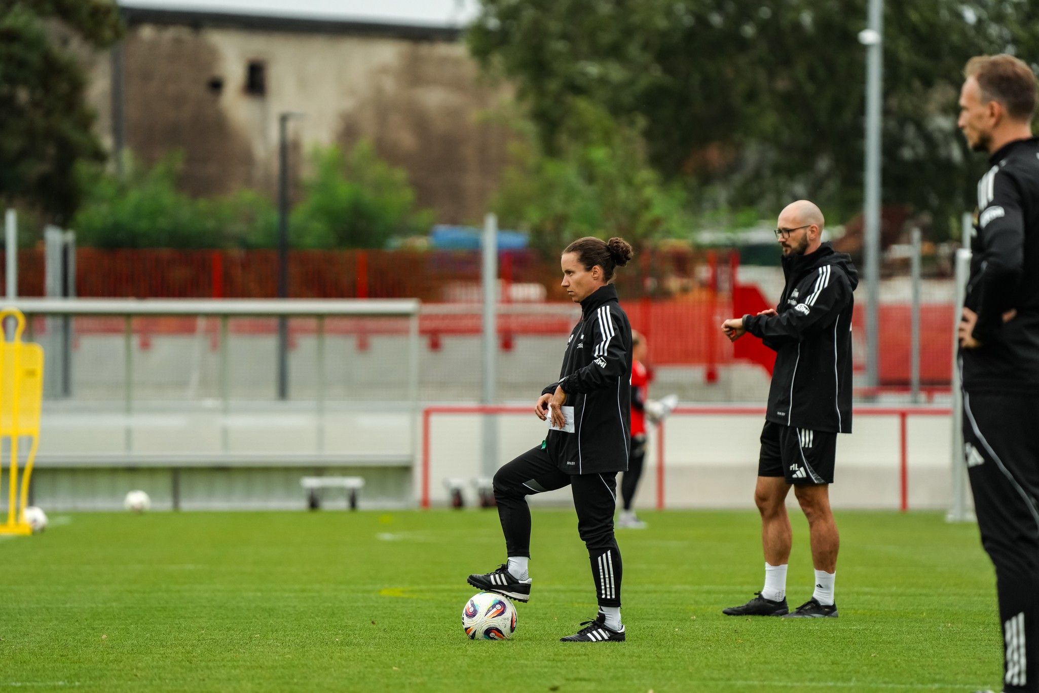 Drei Personen in Trainingsanzügen stehen auf einem Fußballfeld, eine Person bereitet sich auf einen Schuss vor.