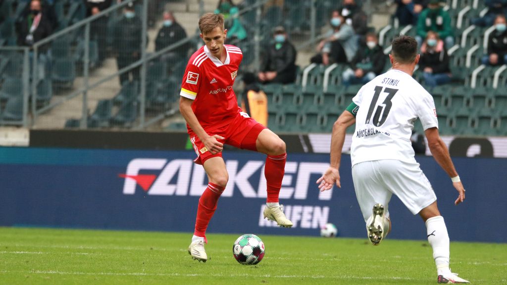 A soccer player in red dribbles the ball while an opponent in white chases him. Spectators in the background.