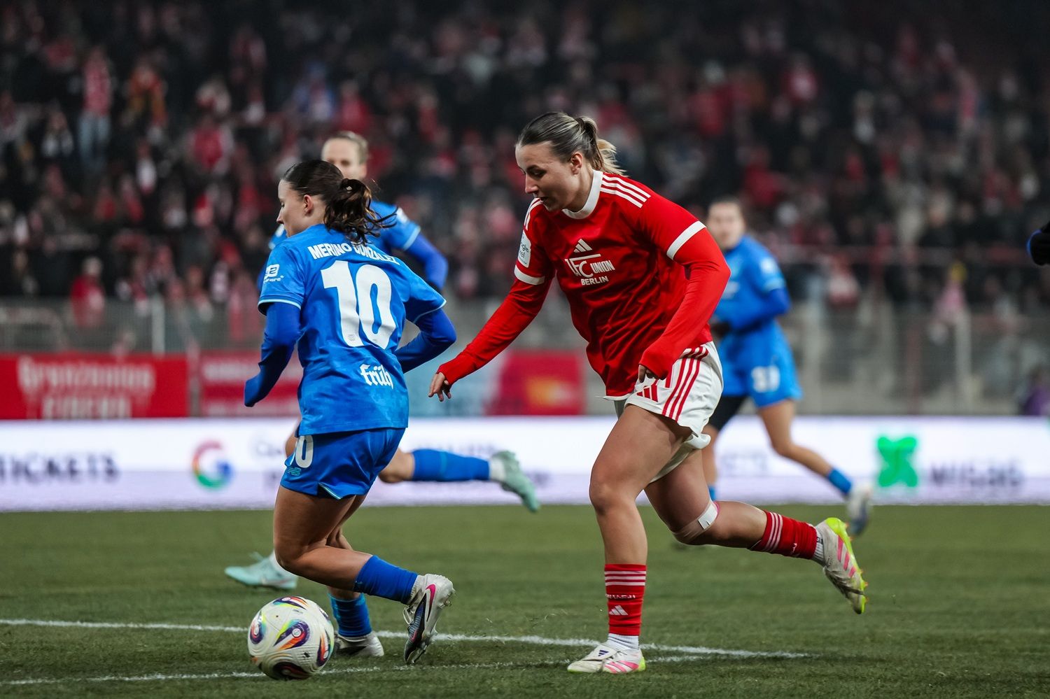 Zwei Fußballspielerinnen in Aktion, eine in blauer und eine in roter Uniform, umgeben von jubelndem Publikum im Hintergrund.