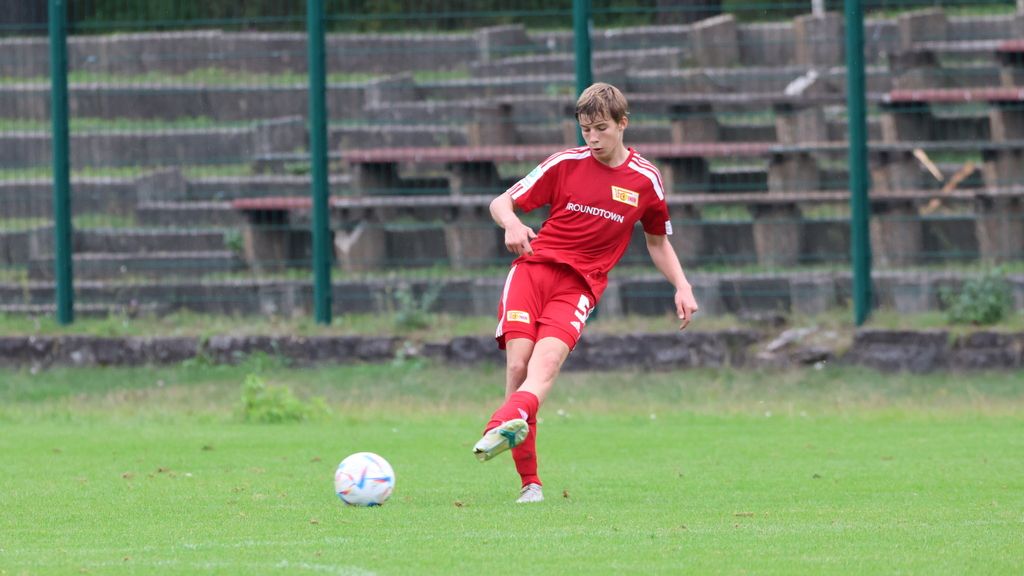 Junger Fußballspieler in rotem Trikot tritt den Ball auf einem grünen Spielfeld mit einer Tribüne im Hintergrund.