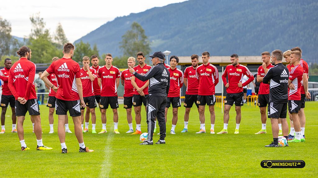 Eine Gruppe von Fußballspielern in roten Trikots versammelt auf einem Trainingsplatz, während ein Trainer Anweisungen gibt.