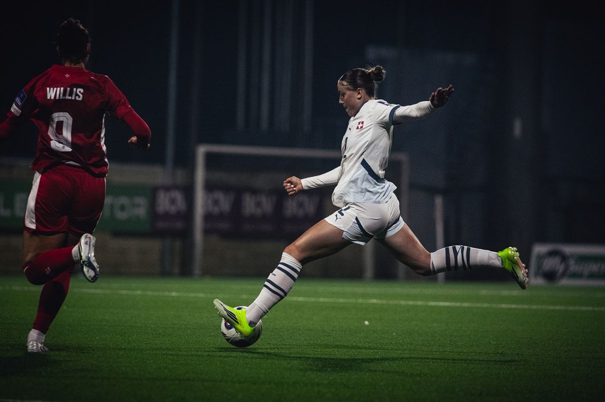Eine Fußballspielerin in weißem Trikot schießt den Ball, während eine Gegnerin in rotem Trikot sich nähert.