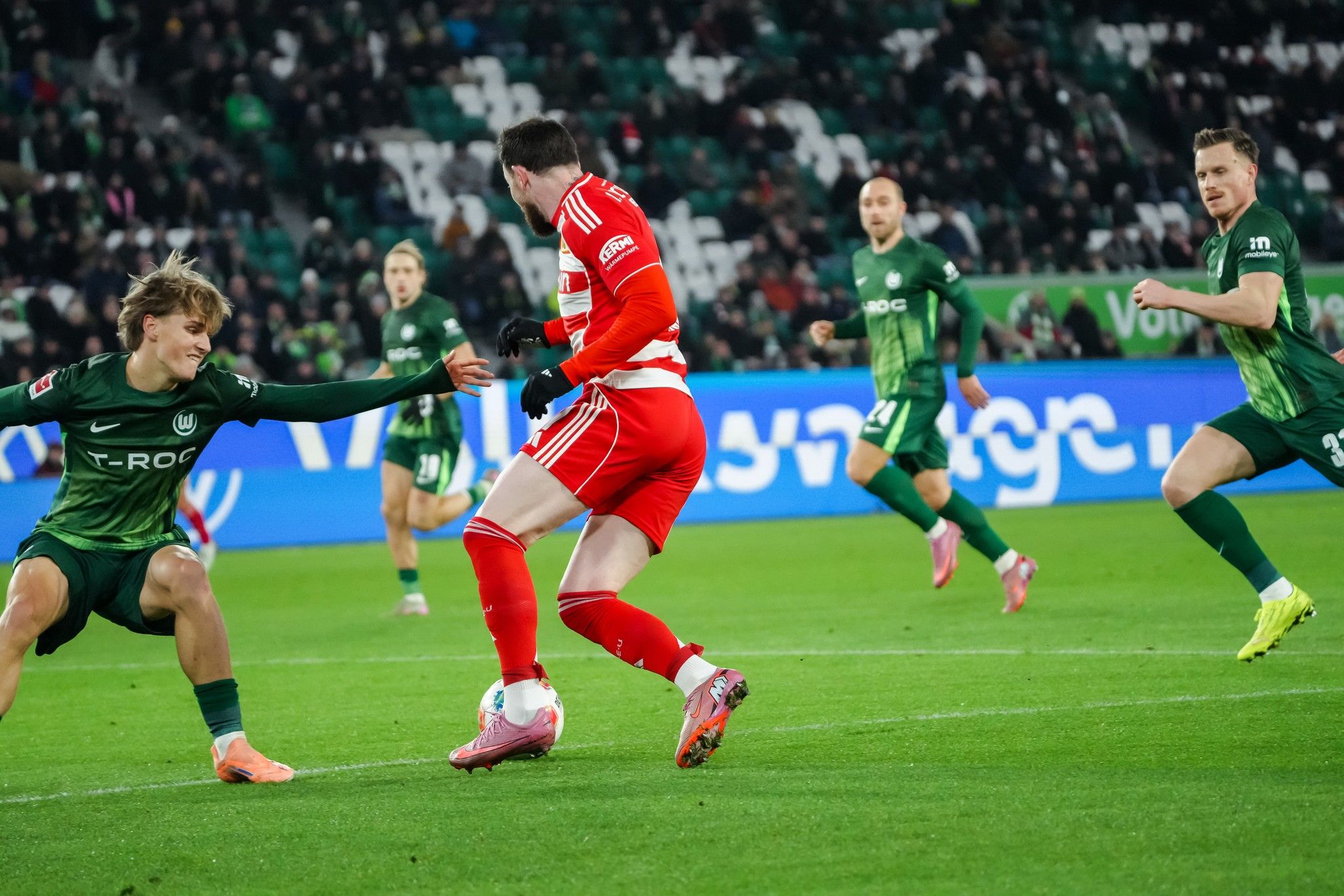 Ein Fußballspieler in roter Ausrüstung wird von einem Gegner in grüner Ausrüstung abgeschirmt, während andere Spieler im Hintergrund zu sehen sind.