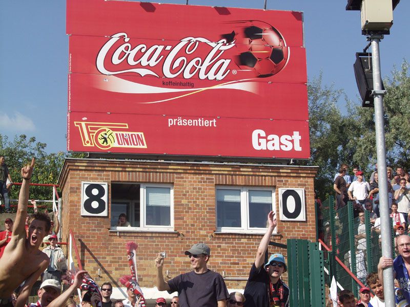 Zahlenschild mit dem Ergebnis 8:0, Werbung für Coca-Cola und Zuschauer im Stadion.