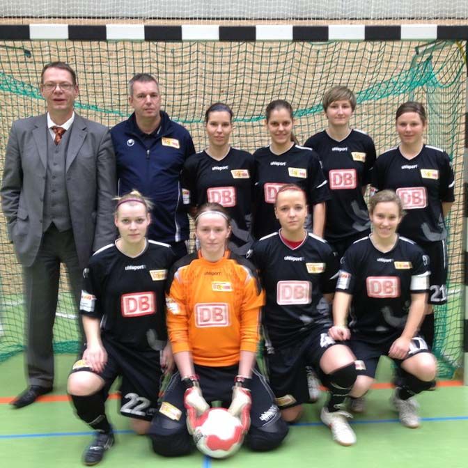Eine Frauenfußballmannschaft in schwarzen Trikots steht zusammen mit zwei Trainern in einem Sporthalle vor einem Fußballtor.