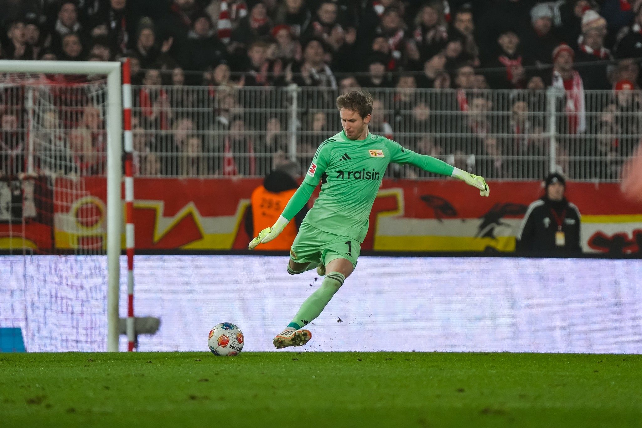 Torhüter in grüner Ausrüstung führt einen Abschlag während eines Fußballspiels durch. Zuschauer im Hintergrund sind sichtbar.