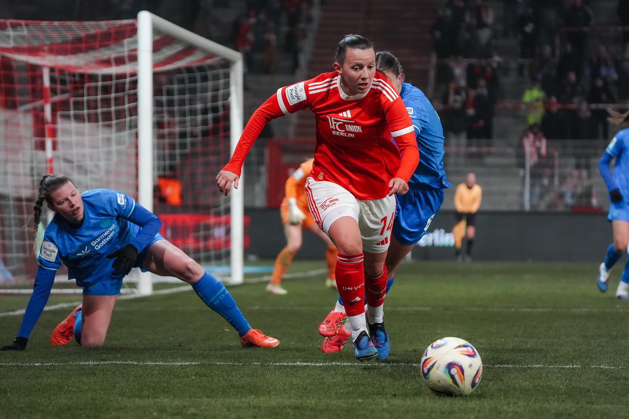 Eine Fußballspielerin in rotem Trikot dribbelt den Ball, während eine Torhüterin versucht, den Ball abzufangen.