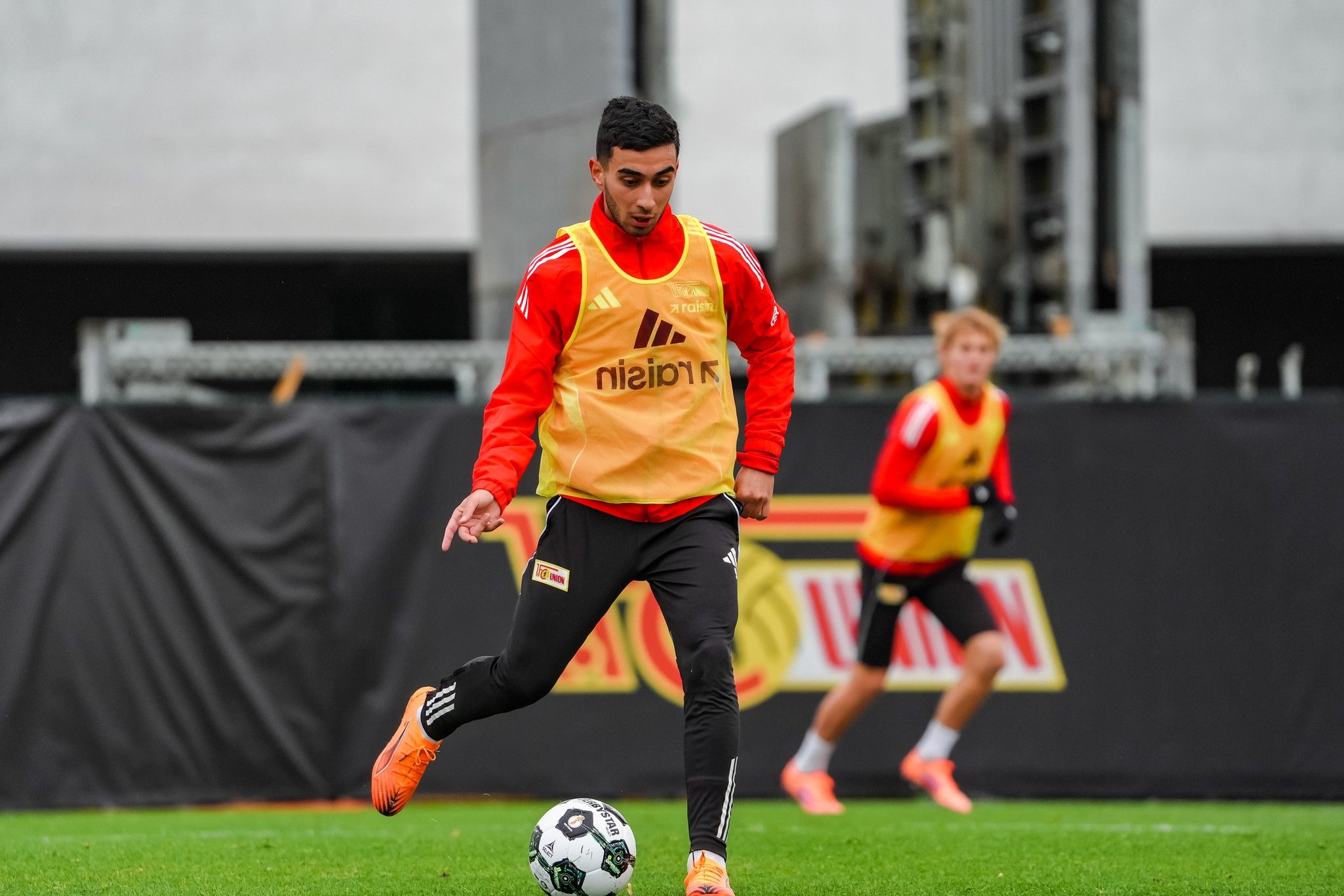 Ein Fußballspieler in Training trägt ein orangefarbens Trikot und spielt den Ball auf einem Sportplatz.