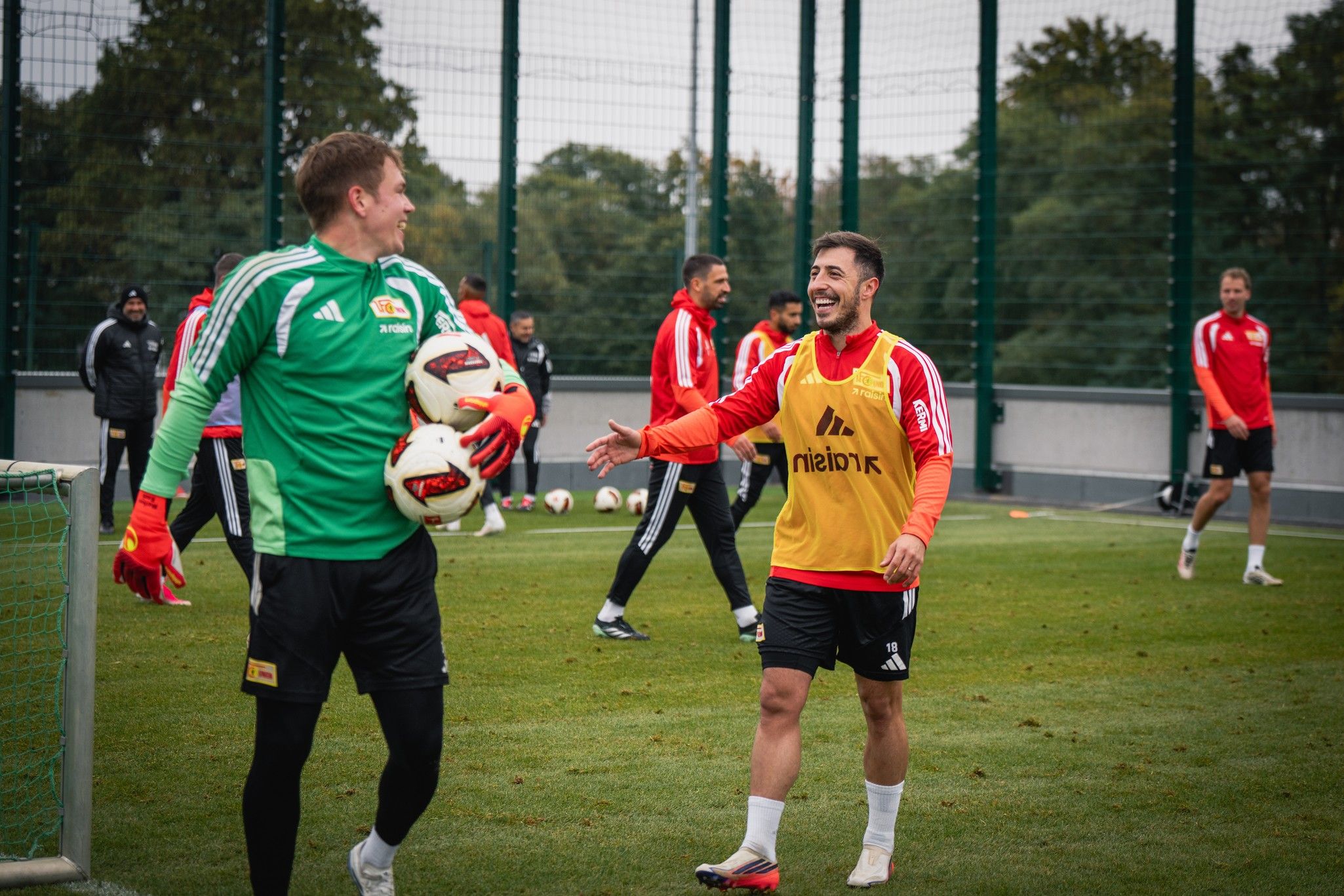Zwei Fußballspieler im Training, einer im Torwart-Trikot, der andere lächelt und gestikuliert auf dem Platz, weitere Spieler im Hintergrund.