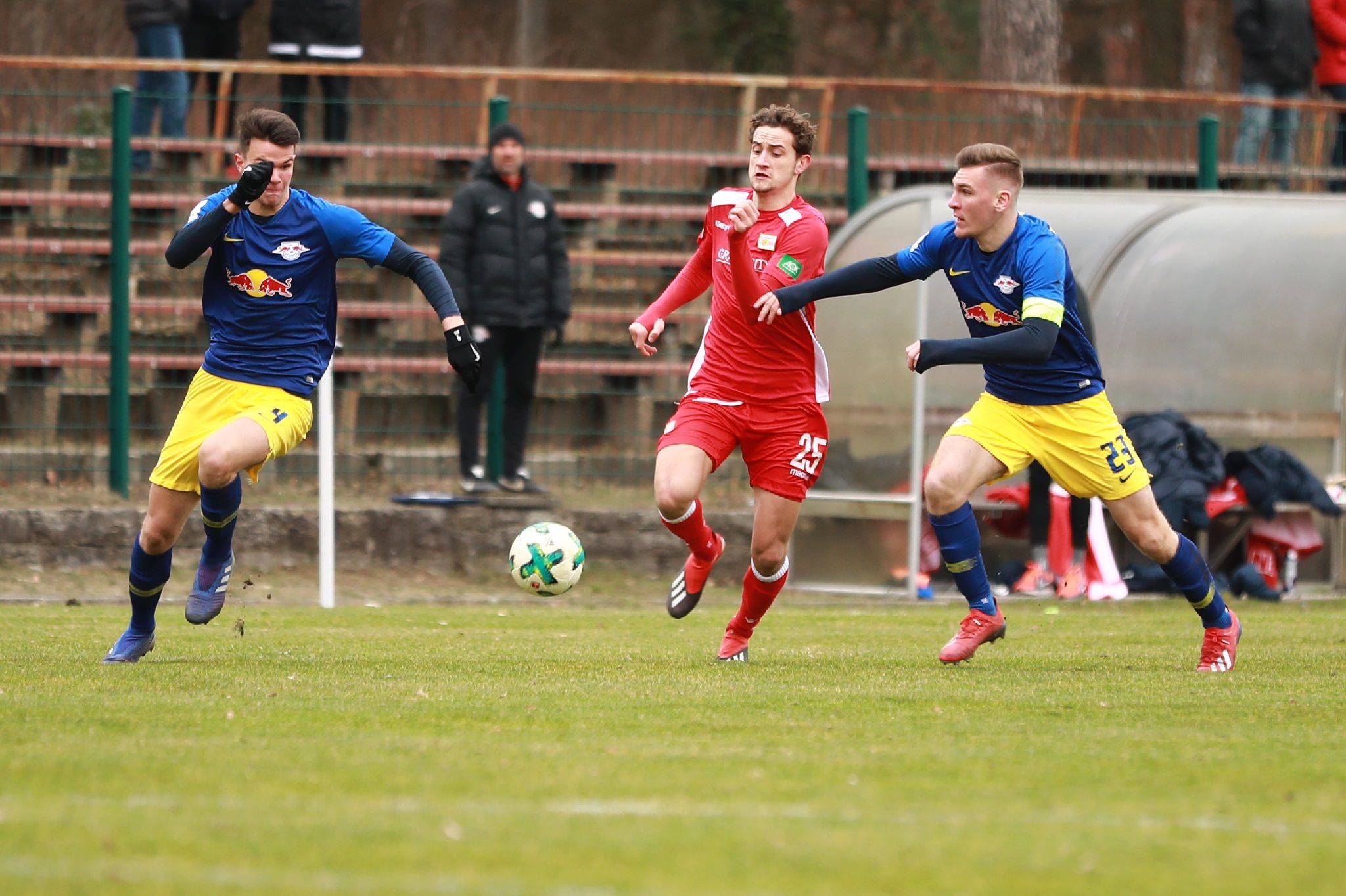 Zwei Fußballspieler in blauen Trikots verfolgen einen Spieler in rotem Trikot, der den Ball in Richtung Tor dribbelt.
