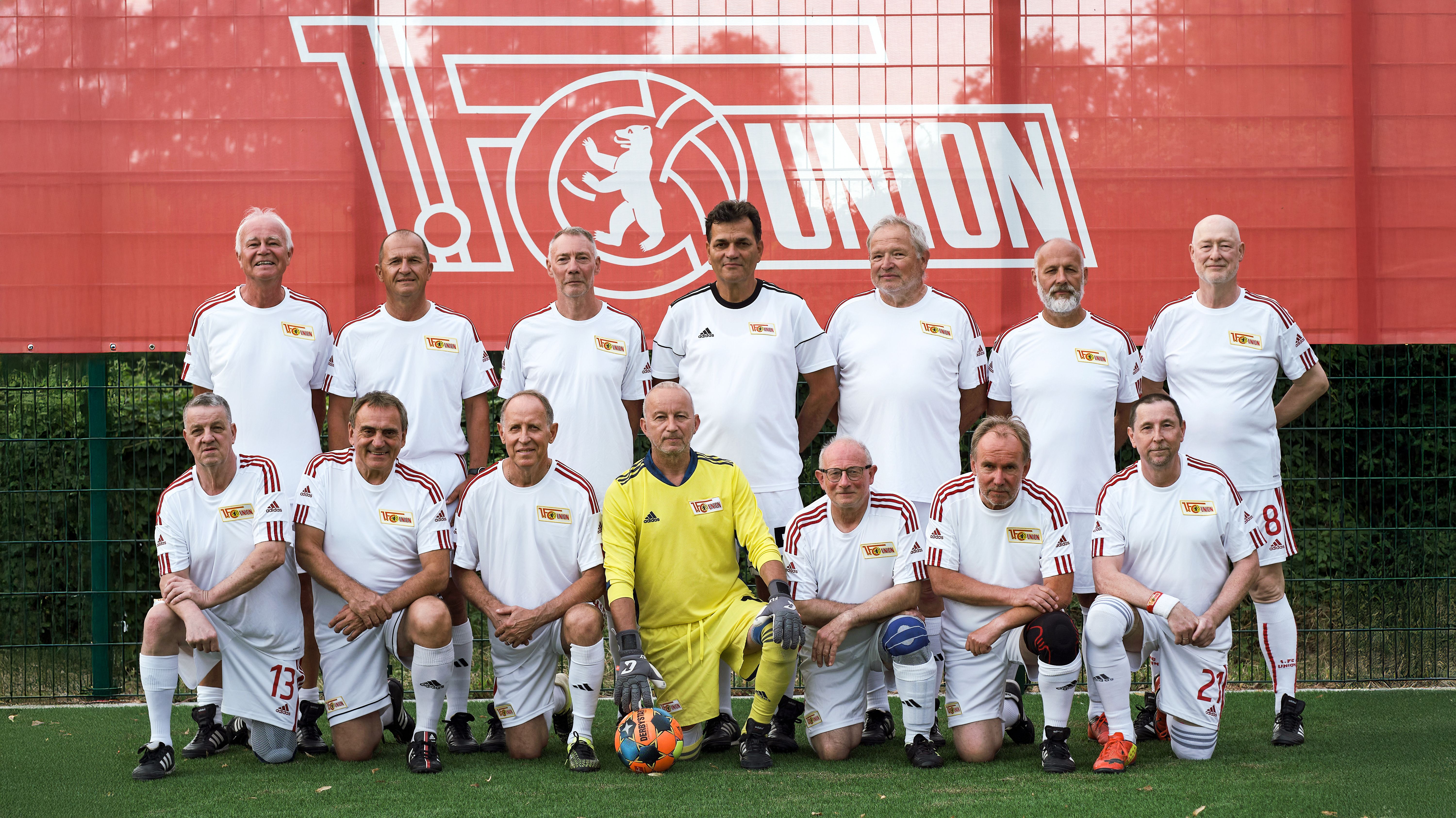 Eine Gruppe von 13 älteren Herren in weißen Trikots steht und kniet auf einem Fußballplatz vor einem rot-weißen Vereinsbanner.
