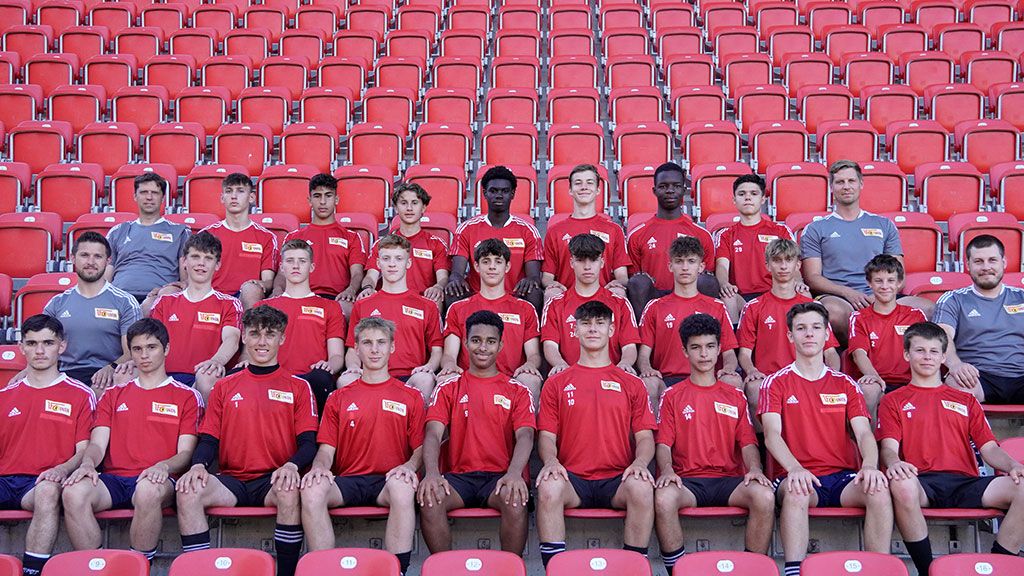 Junge Fußballmannschaft in roten Trikots sitzt in einer Tribüne mit roten Sitzplätzen, Gruppenfoto vor einem sportlichen Hintergrund.