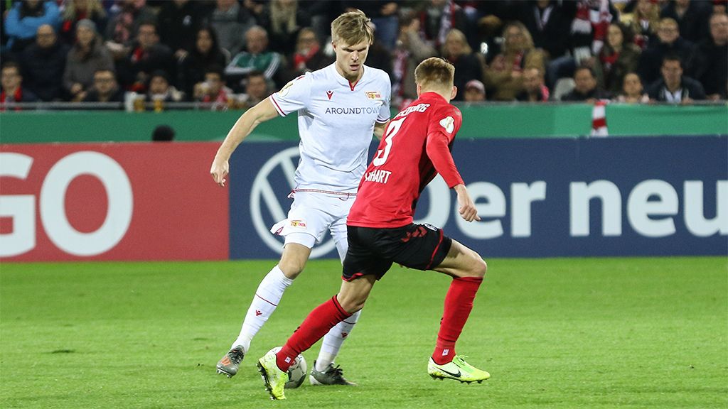 Two football players in action during a game, one dribbling around the other on a stadium field.