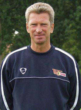 Ein lächelnder Mann in einem dunkelblauen Trainingsanzug steht vor einem unscharfen grünen Hintergrund.