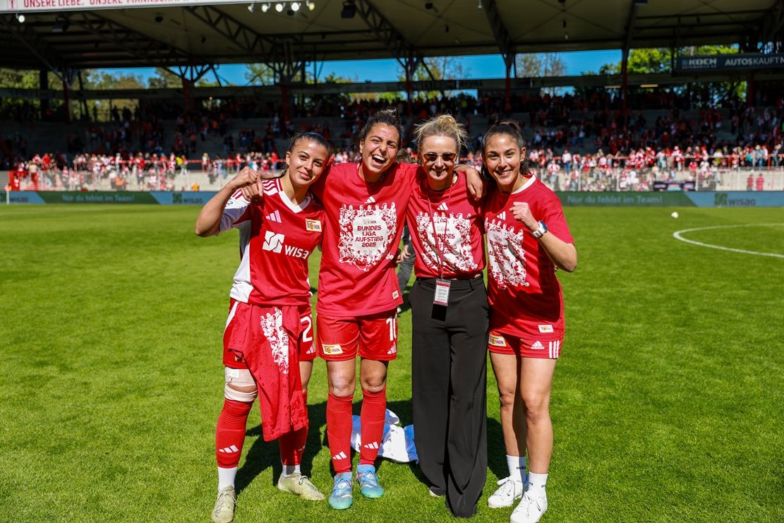 Vier Frauen im roten Fußballtrikot posieren lächelnd auf dem Spielfeld, umgeben von jubelnden Fans im Hintergrund.