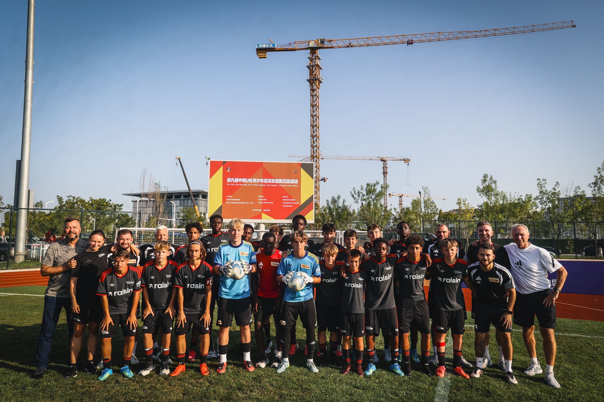 Gruppe von Jugendlichen und Trainern in Fußballtrikots, posierend auf einem Sportplatz mit einem Baukran im Hintergrund.
